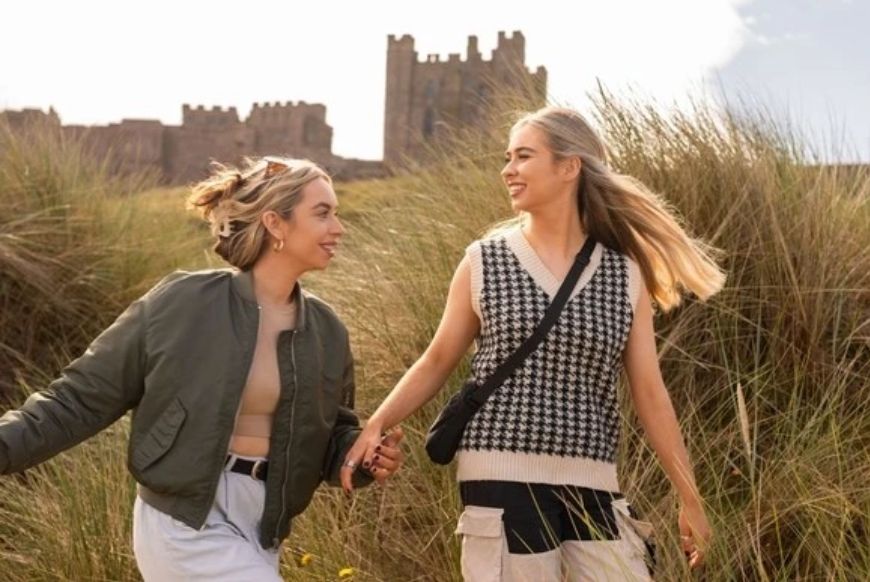 two women holding hands while walking through grass with a castle behind them